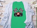 A Garment Graphics green T-shirt with a red racing car and crossed checkered flags is elegantly placed on light wood, paired with a gray cardigan, white high heels, scattered jewelry, and enhanced by white flowers for a charming motorsport-themed display.