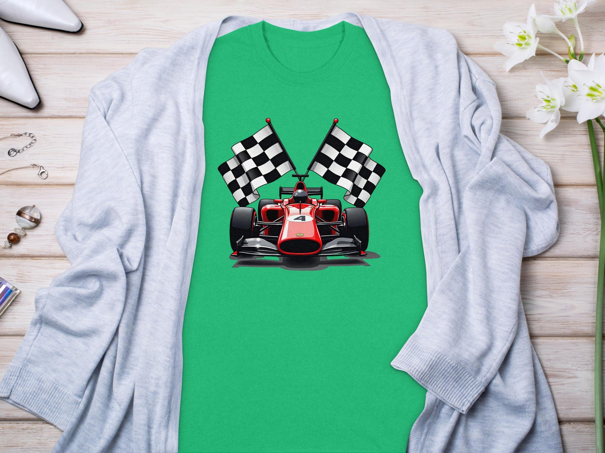 A Garment Graphics green T-shirt with a red racing car and crossed checkered flags is elegantly placed on light wood, paired with a gray cardigan, white high heels, scattered jewelry, and enhanced by white flowers for a charming motorsport-themed display.