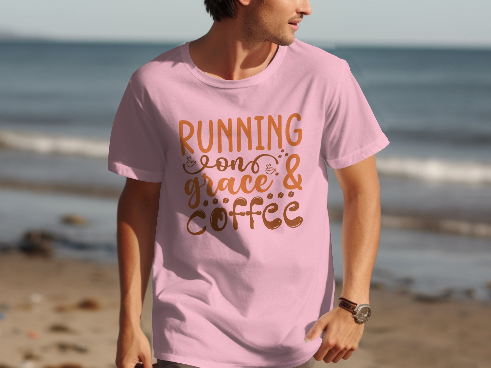 A person walks on the beach in a Garment Graphics T-shirt, showcasing the brown-printed inspirational quote Running on Grace & Coffee against its pink fabric. The tranquil sand and sea backdrop enhances their peaceful stroll.