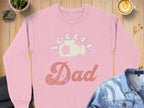 Garment Graphics offers a stylish pink Soccer Dad sweatshirt, featuring a soccer ball in the text. Set on wood with a coffee cup, plant, and denim shirt, this unisex sweatshirt is ideal for any soccer fan.
