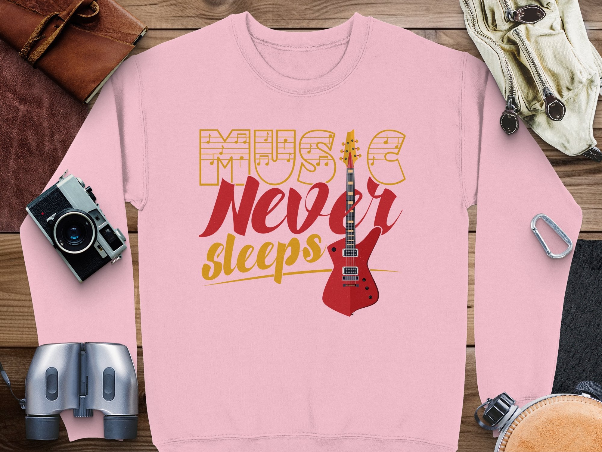 Garment Graphics trendy pink sweatshirt, ideal for music lovers, showcases Music Never Sleeps in bold yellow and red with a vibrant red electric guitar graphic. Its accented by adventurous accessories such as a camera, binoculars, leather gloves, a chic pouch, and climbing carabiners.
