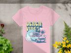 Garment Graphics retro pink t-shirt features a surf-theme design with BEACH VIBES, a vintage car, surfboard, wave, and palm trees. Its stylishly displayed on concrete surrounded by vibrant plants and flowers.