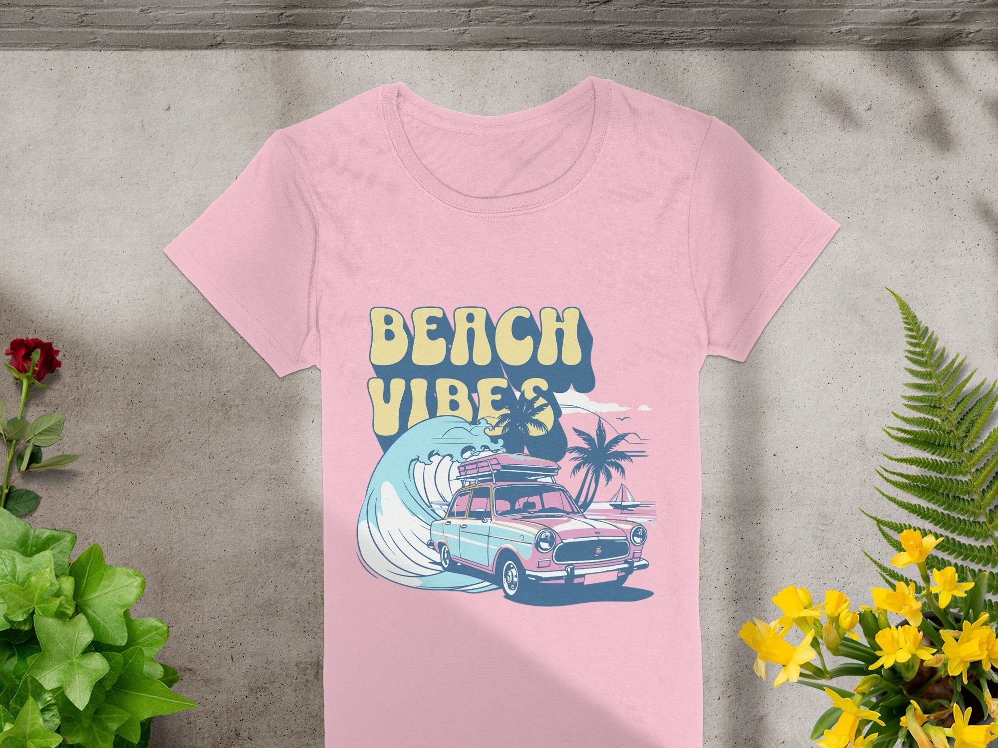 Garment Graphics retro pink t-shirt features a surf-theme design with BEACH VIBES, a vintage car, surfboard, wave, and palm trees. Its stylishly displayed on concrete surrounded by vibrant plants and flowers.