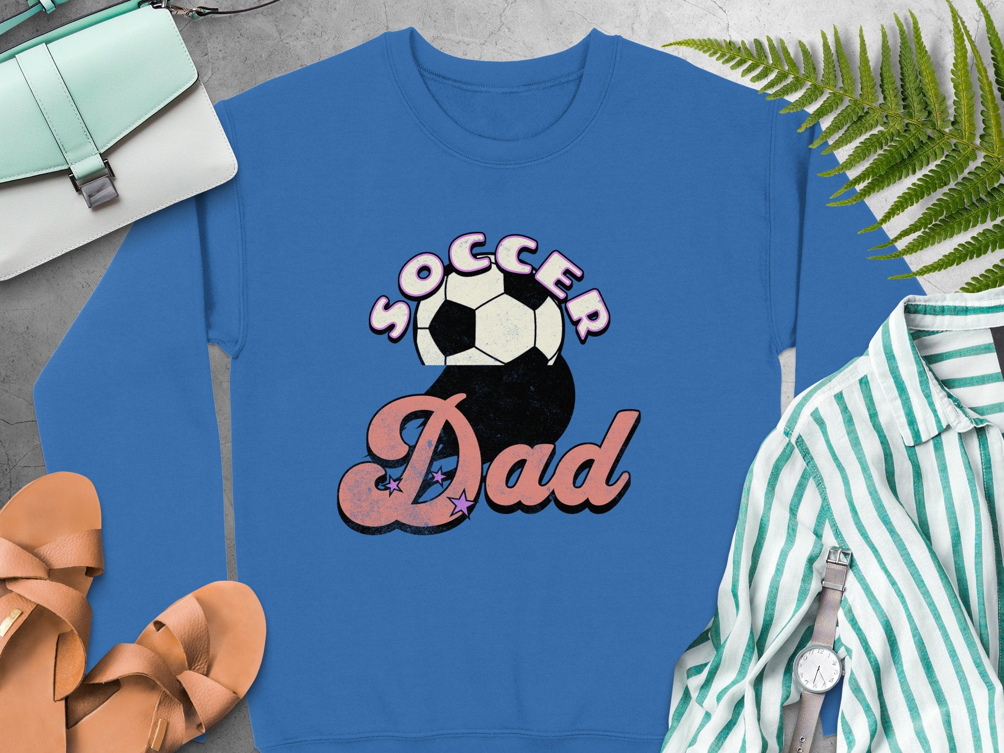 Garment Graphics blue Soccer Dad graphic print sweatshirt with a retro soccer ball design is surrounded by brown sandals, a blue and white striped shirt, a wristwatch, a fern leaf, and a light blue purse on a gray surface.