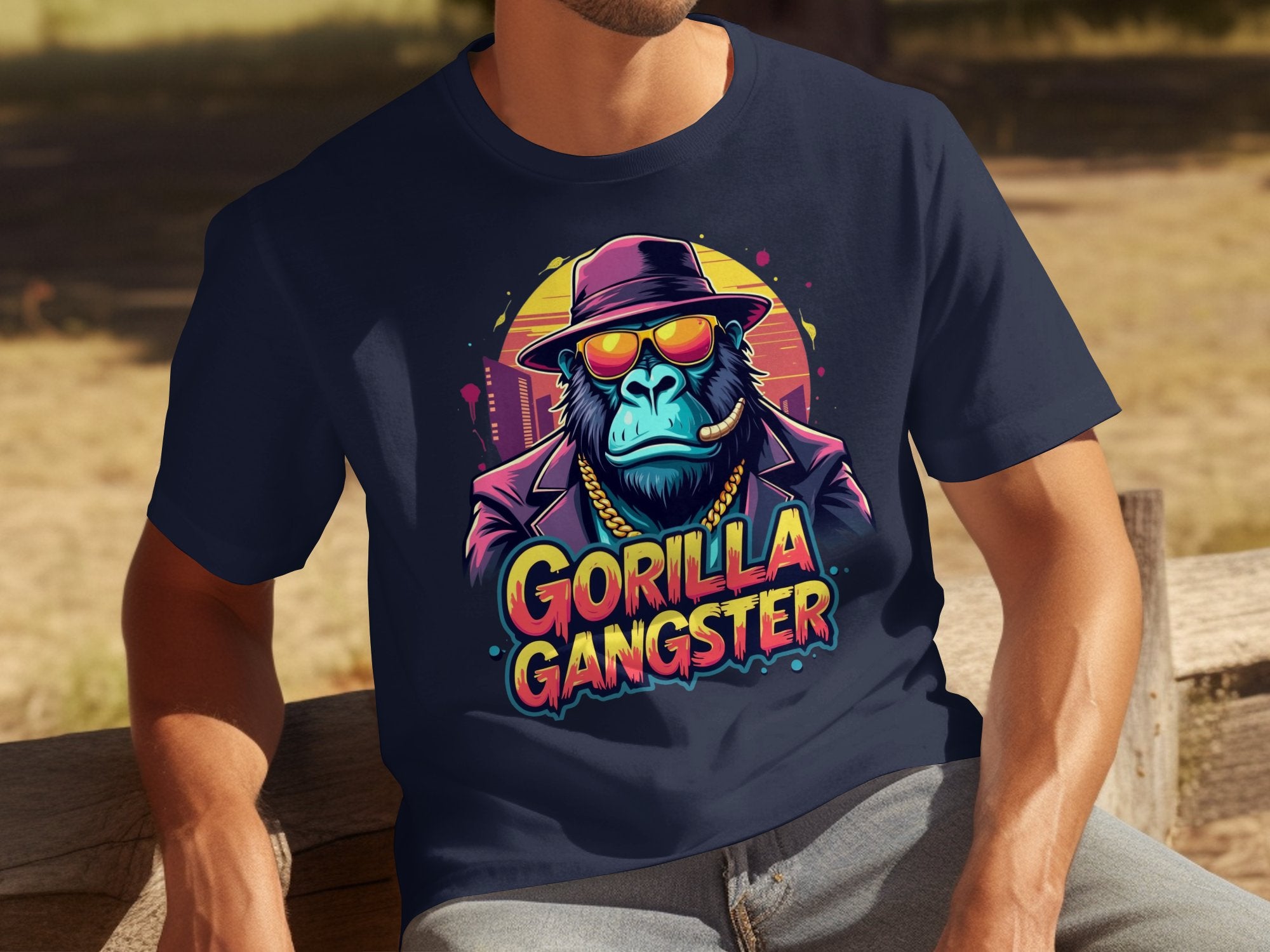 Seated on a wooden bench, a person makes a bold statement in a Garment Graphics navy t-shirt. It has a colorful graphic of a Gorilla Gangster wearing sunglasses, a hat, and chain, with GORILLA GANGSTER boldly printed below.