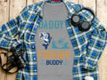 A gray T-shirt from Garment Graphics, featuring Daddys Fishing Buddy text with a fish and fisherman on a boat graphic, lies on a wooden surface. Ideal for outdoor lovers, its paired with a blue plaid shirt, headphones, a black camera, and a white tumbler.