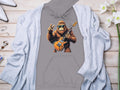 Garment Graphics gray hoodie showcases a cartoon gorilla in sunglasses, holding an electric guitar and making a peace sign—ideal for music lovers. Laid on light wood, its surrounded by a gray cardigan, white flowers, jewelry, and white high heels.