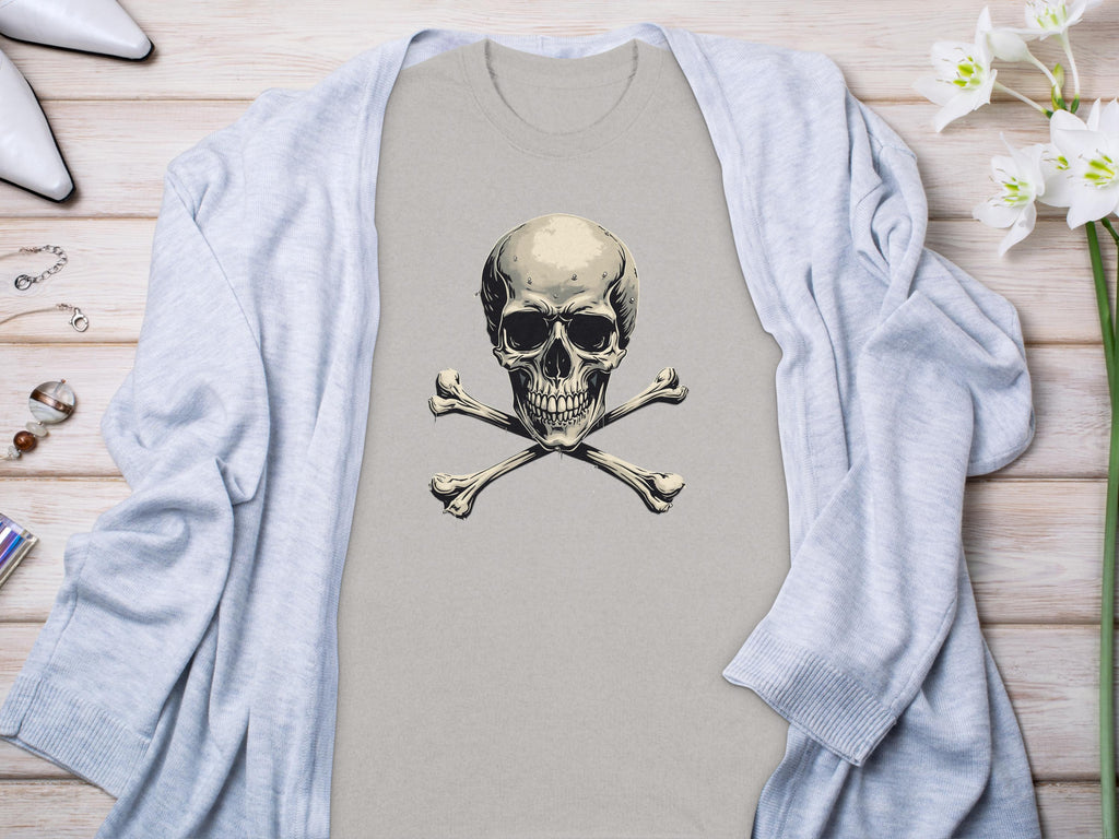 A beige skull and crossbones graphic t-shirt by Garment Graphics is styled on a light wooden surface. A gray cardigan drapes over it, enhancing the chic look. White high-heeled shoes, a bracelet, ring, and white flowers complete the fashionable ensemble.