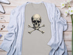 A beige skull and crossbones graphic t-shirt by Garment Graphics is styled on a light wooden surface. A gray cardigan drapes over it, enhancing the chic look. White high-heeled shoes, a bracelet, ring, and white flowers complete the fashionable ensemble.