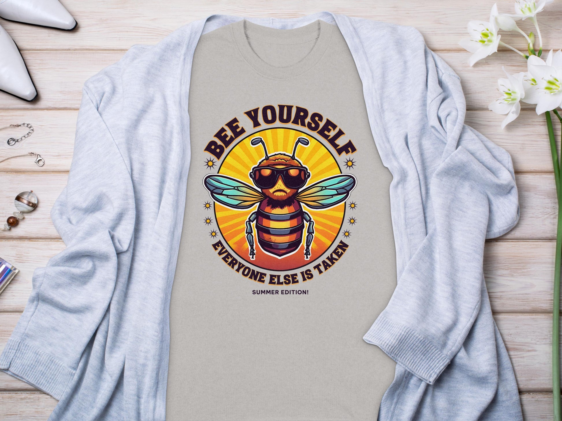 A light gray T-shirt from Garment Graphics showcases a hip bee in sunglasses with the phrase Bee Yourself, Every One Else Is Taken, Summer Edition! Ideal for family sizing, its stylishly paired with a cardigan, perfume, jewelry, and artfully arranged white flowers.