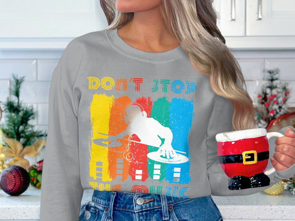 A person in a Garment Graphics gray sweatshirt with a vibrant DJ design and Dont Stop the Music text, perfect for music lovers, holds a Santa-themed mug. The white kitchen behind, adorned with festive decorations, adds to the cozy holiday ambiance.