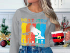 A person in a Garment Graphics gray sweatshirt with a vibrant DJ design and Dont Stop the Music text, perfect for music lovers, holds a Santa-themed mug. The white kitchen behind, adorned with festive decorations, adds to the cozy holiday ambiance.