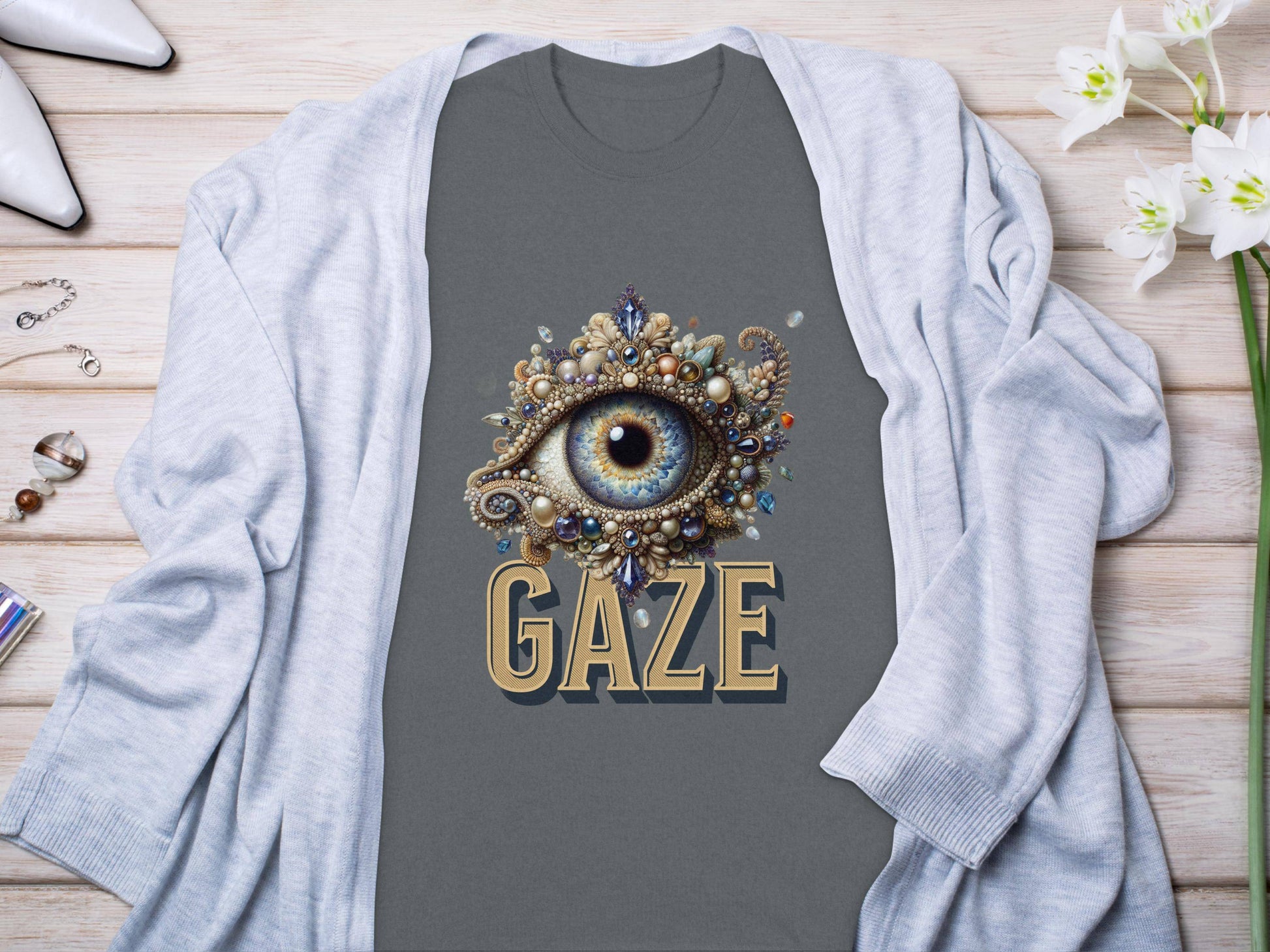 A captivating eye illustration shirt from Garment Graphics, in dark gray, displays the word Gaze. Its styled on a light wooden surface with a light gray cardigan, white shoes, jewelry, and white flowers.