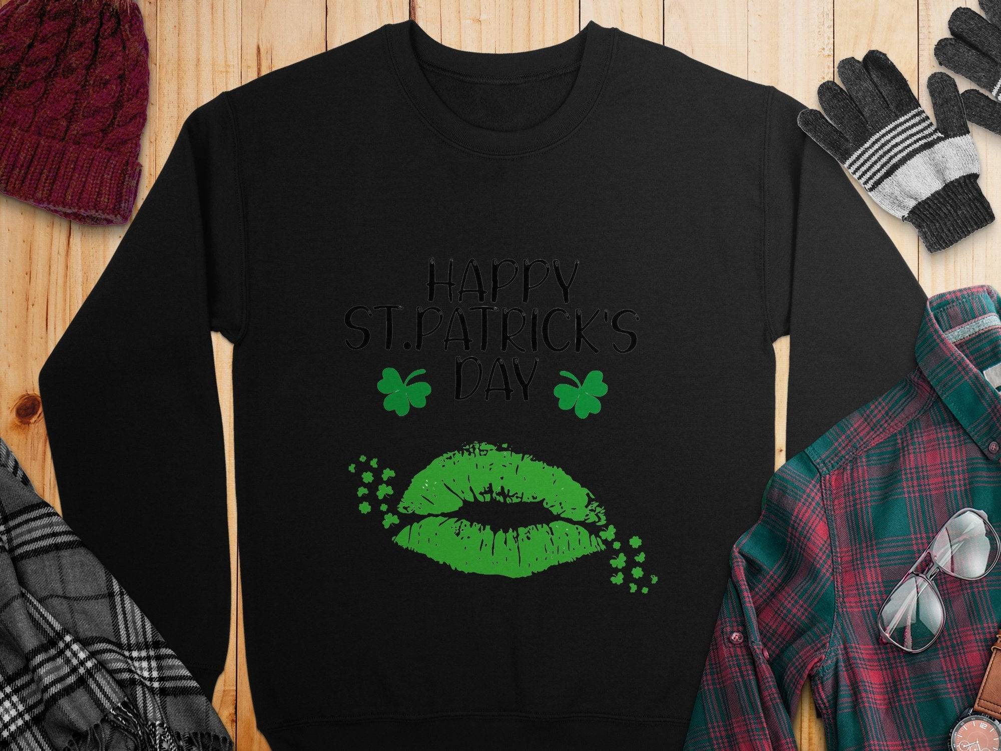 A black Garment Graphics sweatshirt showcases Happy St. Patricks Day in script with green lips and shamrocks, capturing the festive clover kiss vibe. Its stylishly matched on a wooden surface with a red knit hat, black and gray gloves, a plaid green shirt, and a gray plaid scarf.