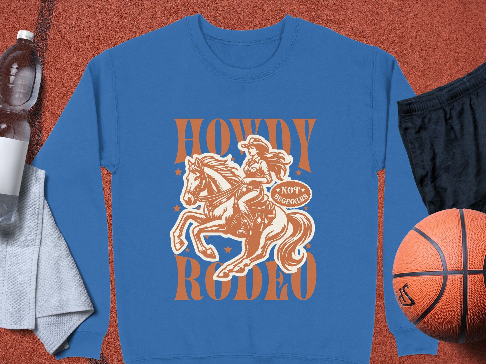 A unisex blue graphic print sweatshirt by Garment Graphics features a cowboy riding a horse with Howdy Rodeo text, holding a Not to Beginners shield, surrounded by sports items including a basketball, black shorts, towel, and water bottle.