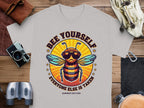 The Garment Graphics gray t-shirt features a bee in sunglasses and the playful text BEE YOURSELF EVERYONE ELSE IS TAKEN above and below. Ideal for fans of unique Every One Else Is Taken T-Shirts, it appears with a camera, binoculars, wallet, and sandals on wooden planks.