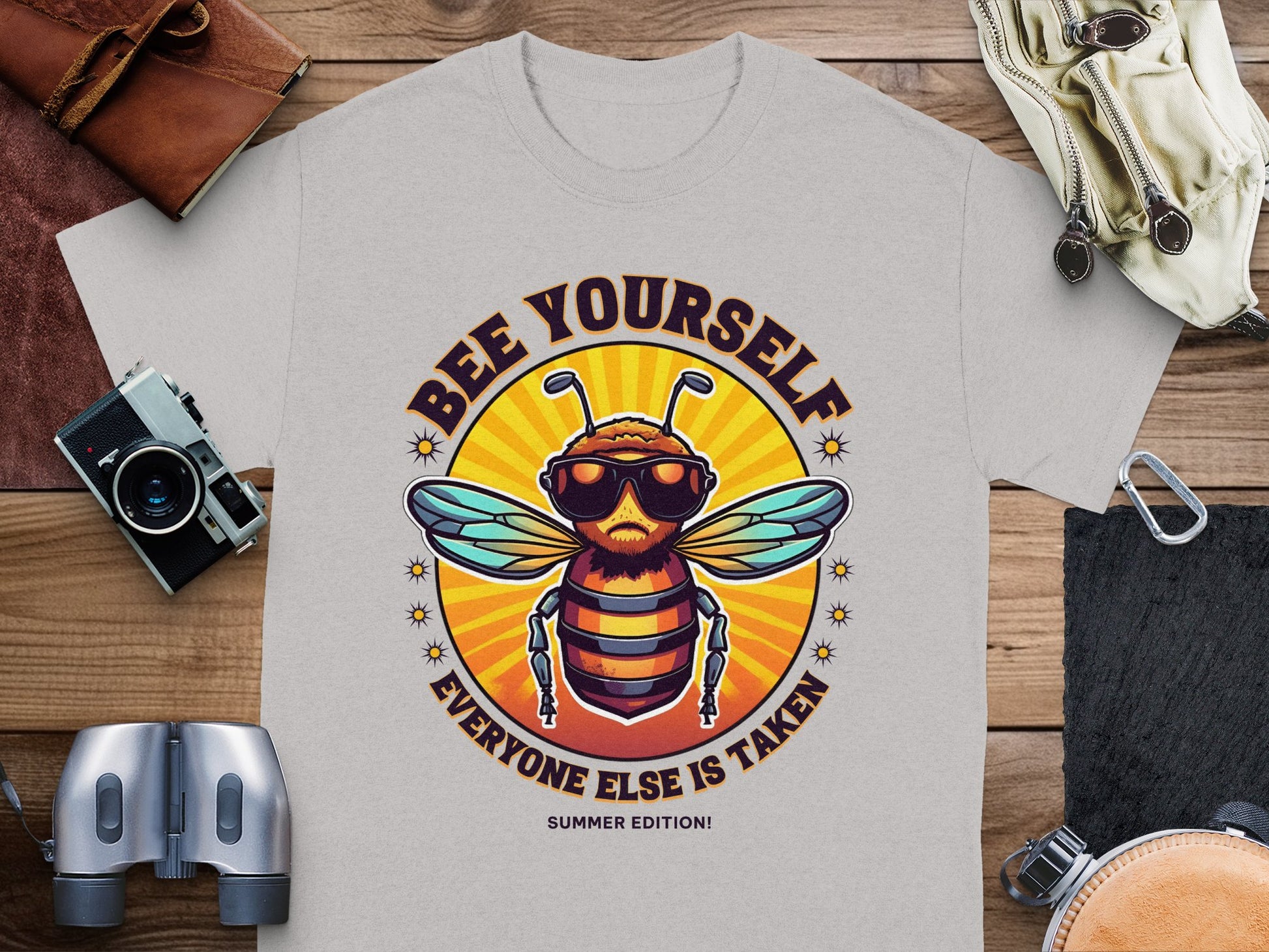 The Garment Graphics gray t-shirt features a bee in sunglasses and the playful text BEE YOURSELF EVERYONE ELSE IS TAKEN above and below. Ideal for fans of unique Every One Else Is Taken T-Shirts, it appears with a camera, binoculars, wallet, and sandals on wooden planks.