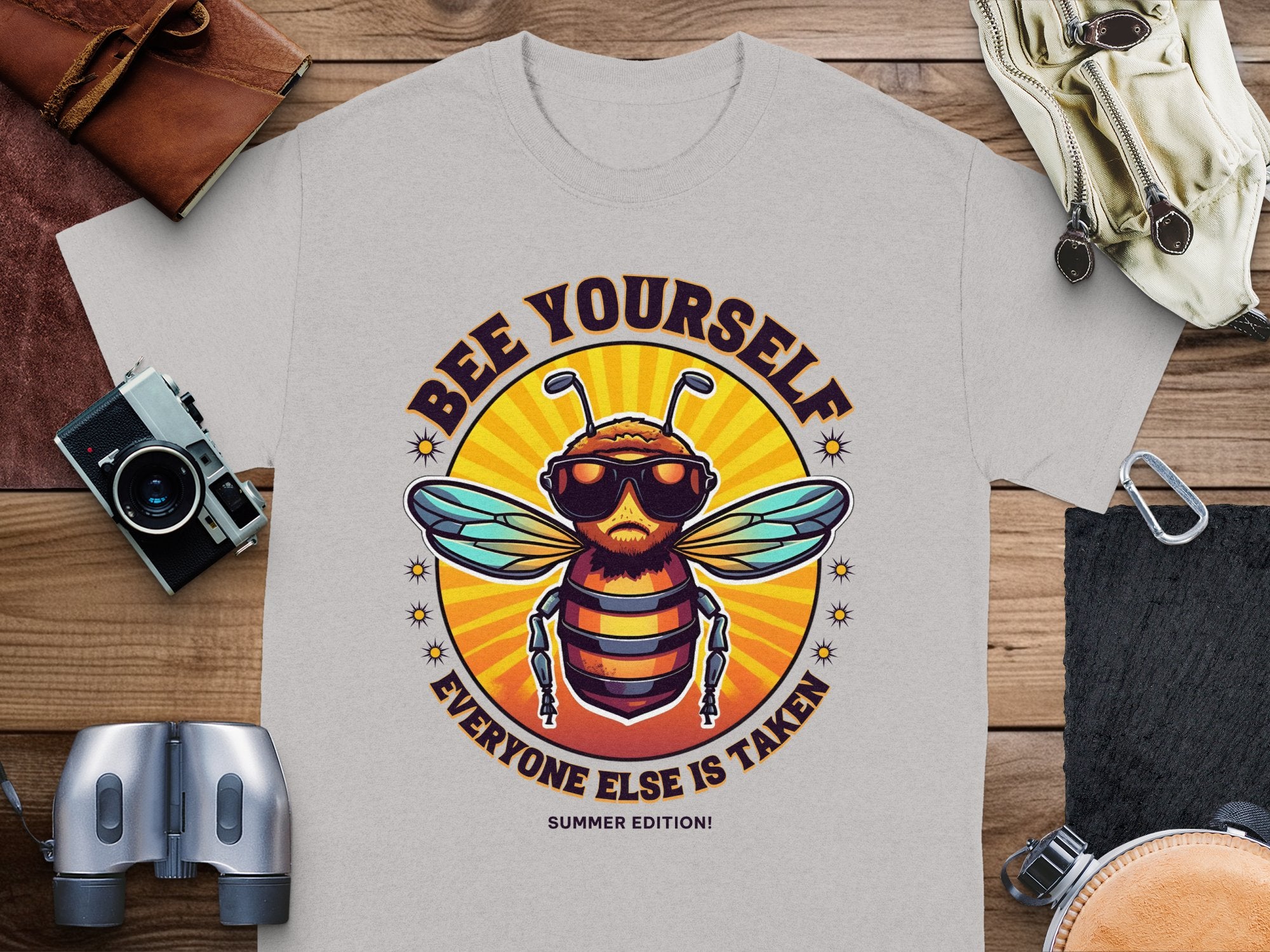 The Garment Graphics gray t-shirt features a bee in sunglasses and the playful text BEE YOURSELF EVERYONE ELSE IS TAKEN above and below. Ideal for fans of unique Every One Else Is Taken T-Shirts, it appears with a camera, binoculars, wallet, and sandals on wooden planks.