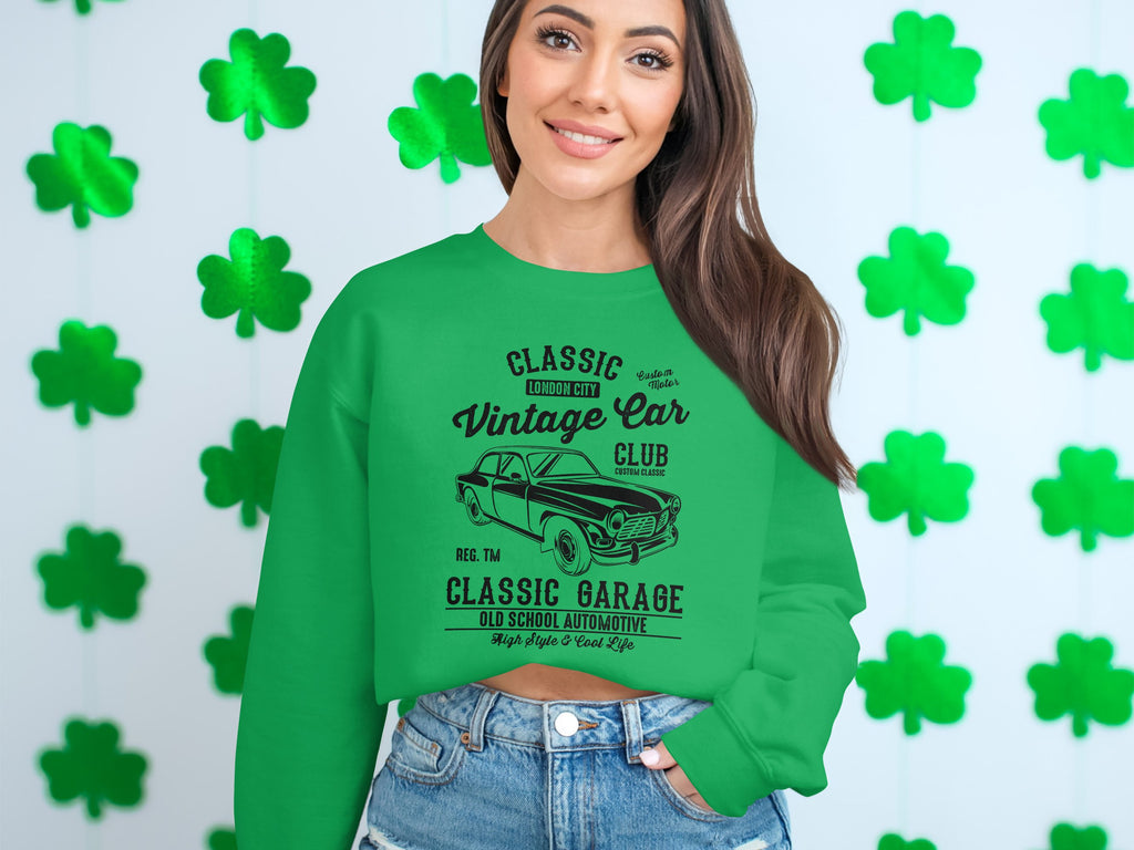 In front of a shamrock backdrop, a person wears a Garment Graphics green sweatshirt featuring a vintage car and Classic Garage text, perfectly embodying classic London city vibes.
