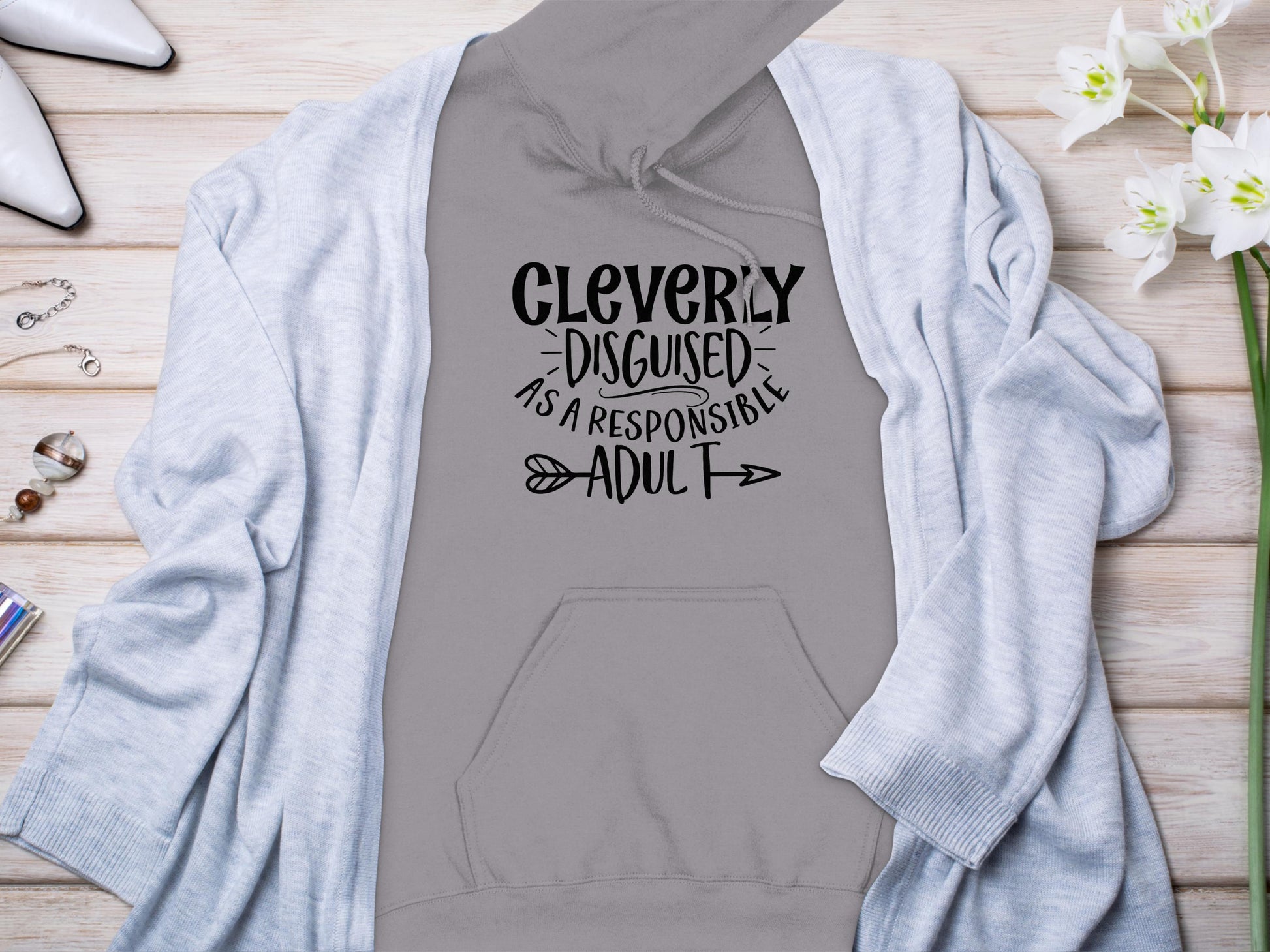 The Garment Graphics gray hoodie, featuring Cleverly disguised as a responsible adult in black font, is perfect for those seeking Responsible Adult Hoodies. Styled on wood, it pairs effortlessly with a light gray cardigan, white flowers, and shoes for a casual vibe.