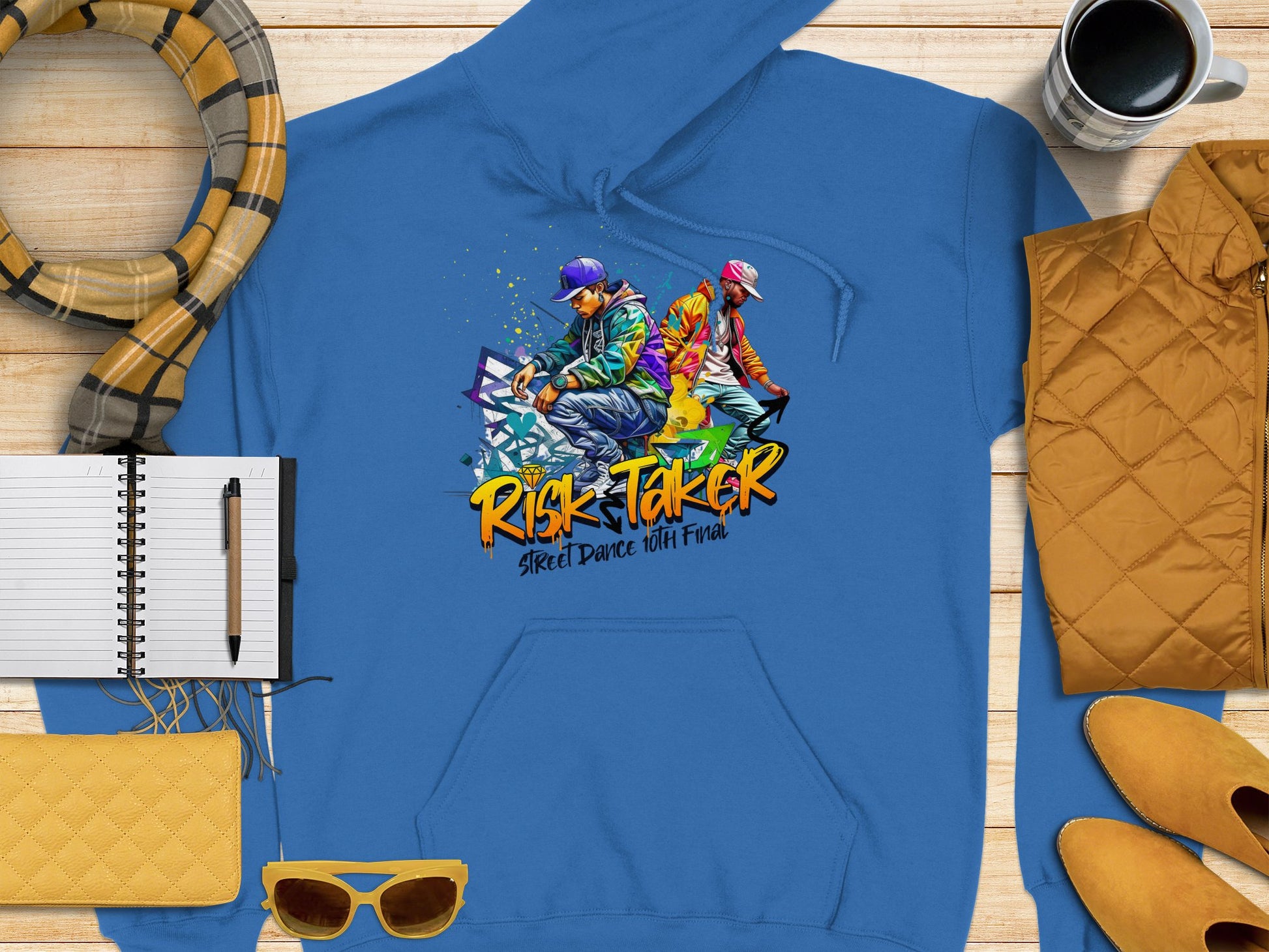 Garment Graphics blue hoodie showcases a graphic of two vividly dressed street dancers and graffiti art, celebrating Risk Taker Street Dance 10th Final. Perfectly complete the edgy street style with a notebook, pen, coffee cup, scarf, yellow sunglasses, and jacket.