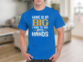 A person wearing a classic fit blue graphic tee by Garment Graphics, featuring the humorous slogan MINE IS SO BIG I HAVE TO USE BOTH HANDS, stands in a room with blurred furnishings in the background.