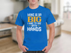 A person wearing a classic fit blue graphic tee by Garment Graphics, featuring the humorous slogan MINE IS SO BIG I HAVE TO USE BOTH HANDS, stands in a room with blurred furnishings in the background.