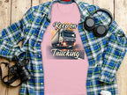 The Garment Graphics pink T-shirt boasts a Keep on Trucking print featuring a truck design, ideal for truck lovers. Its displayed with a plaid shirt, black headphones, a camera, and a white cup on the wooden surface.