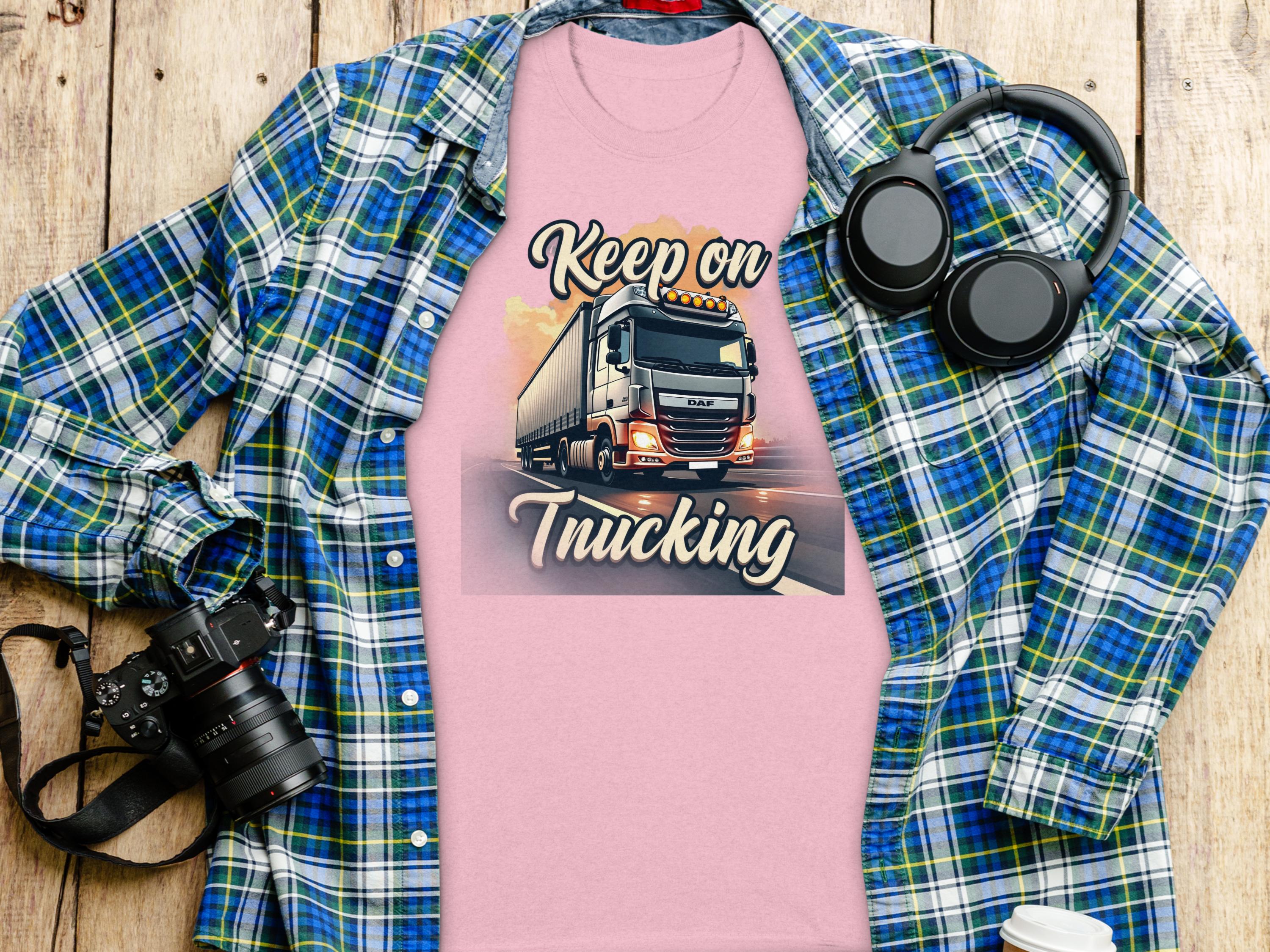 The Garment Graphics pink T-shirt boasts a Keep on Trucking print featuring a truck design, ideal for truck lovers. Its displayed with a plaid shirt, black headphones, a camera, and a white cup on the wooden surface.