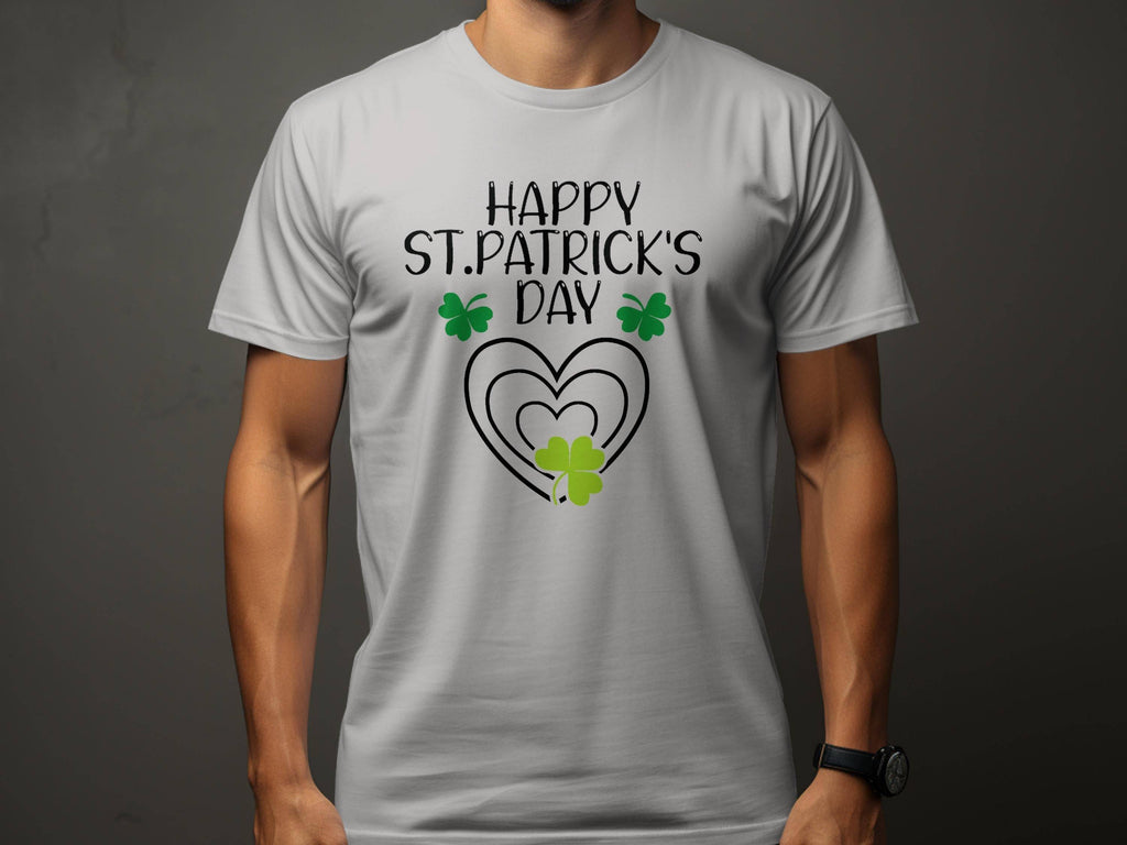 A man wears a Garment Graphics festive flair tee featuring Happy St. Patricks Day in black, with three green shamrocks and a heart integrated into the text, all on a neutral gray background.