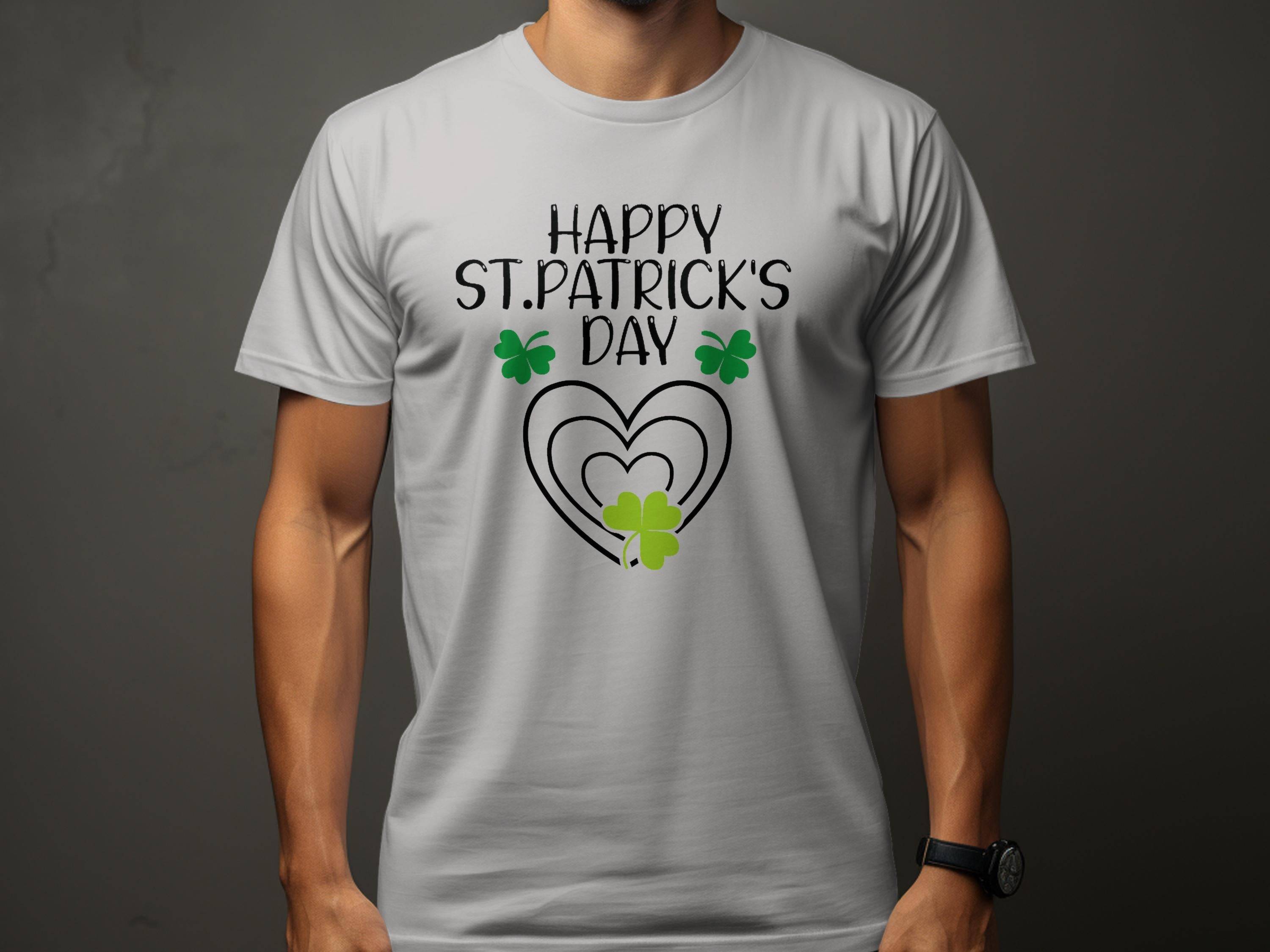 A man wears a Garment Graphics festive flair tee featuring Happy St. Patricks Day in black, with three green shamrocks and a heart integrated into the text, all on a neutral gray background.