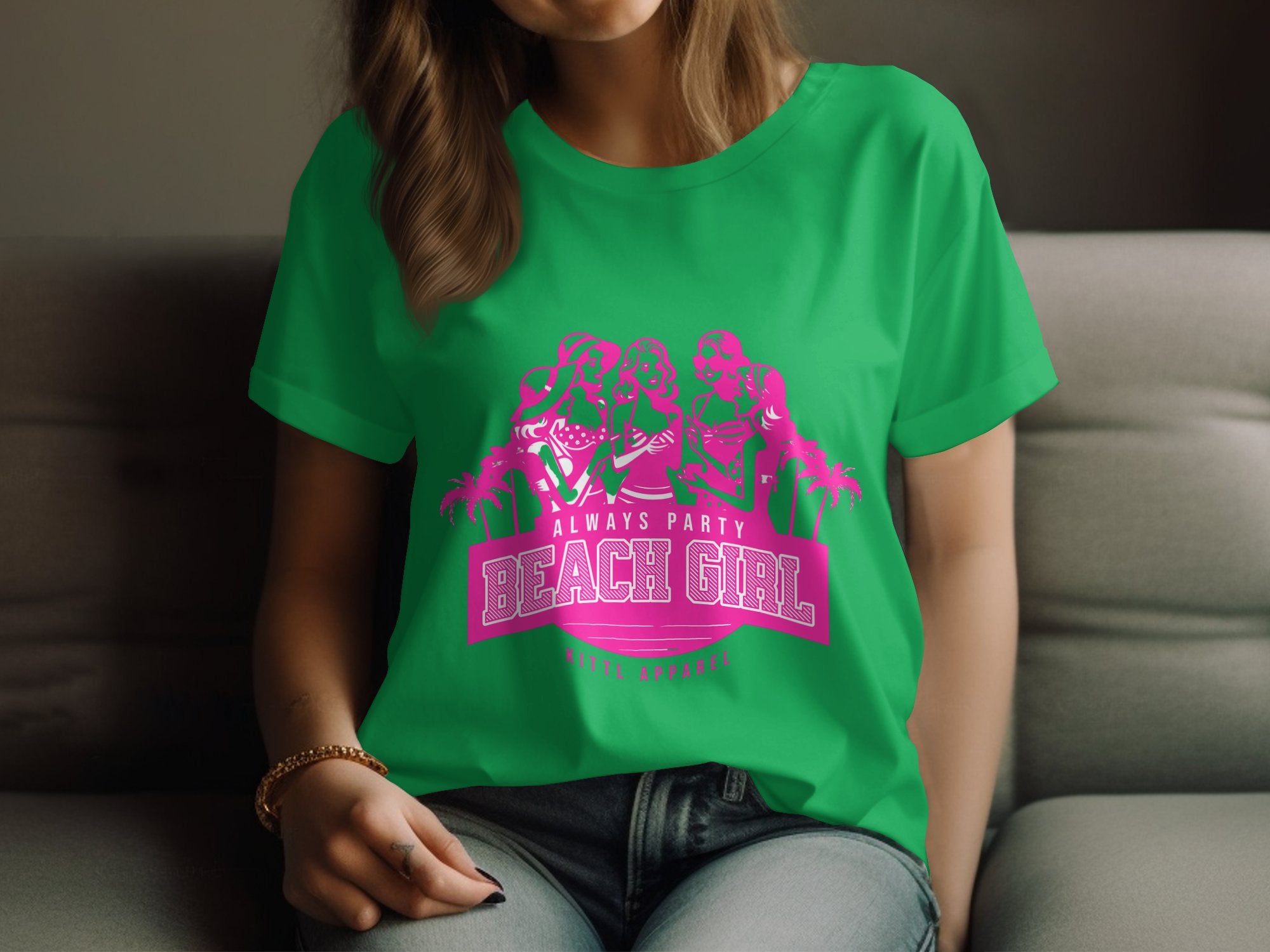 A person lounges on a sofa, wearing a Garment Graphics green T-shirt. The pink graphic shows three styled figures above the text Always Party Beach Girl, with palm tree silhouettes. They gently hold the hem of this Womens Semi-Fitted Tee.