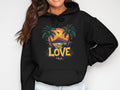 A person sports a black graphic hoodie by Garment Graphics, displaying a tropical sunset with palm trees and a flying bird. One is in bright yellow and Love in vivid green. With one hand on their hip and face partially visible, they exude confidence and distinctive style.