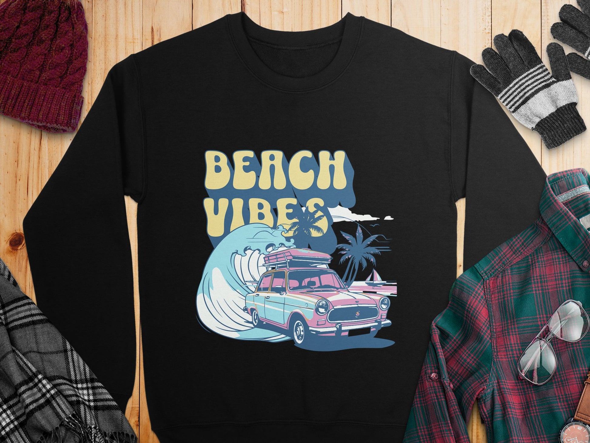 Garment Graphics presents a retro black sweatshirt featuring a pink and blue classic car with surfboards, riding waves with Beach Vibes text. Perfect for cozy beach fun, pair it with winter accessories like beanies, gloves, and plaid shirts.