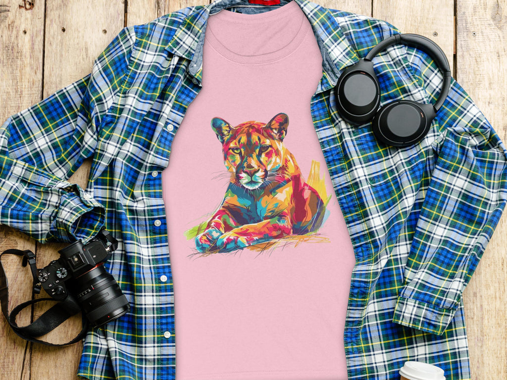 A Garment Graphics pink t-shirt with a vibrant puma print is laid on wood, surrounded by a blue and green plaid shirt, black headphones, a camera, and a coffee cup.