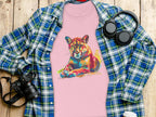 A Garment Graphics pink t-shirt with a vibrant puma print is laid on wood, surrounded by a blue and green plaid shirt, black headphones, a camera, and a coffee cup.
