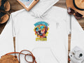 Garment Graphics white hoodie showcases a vibrant dog graphic in sunglasses and hat, with Pawsitively Stylish text. Perfect against wood surfaces, it pairs well with sandals, a notebook, camera, and straw hat with shades for a complete look.