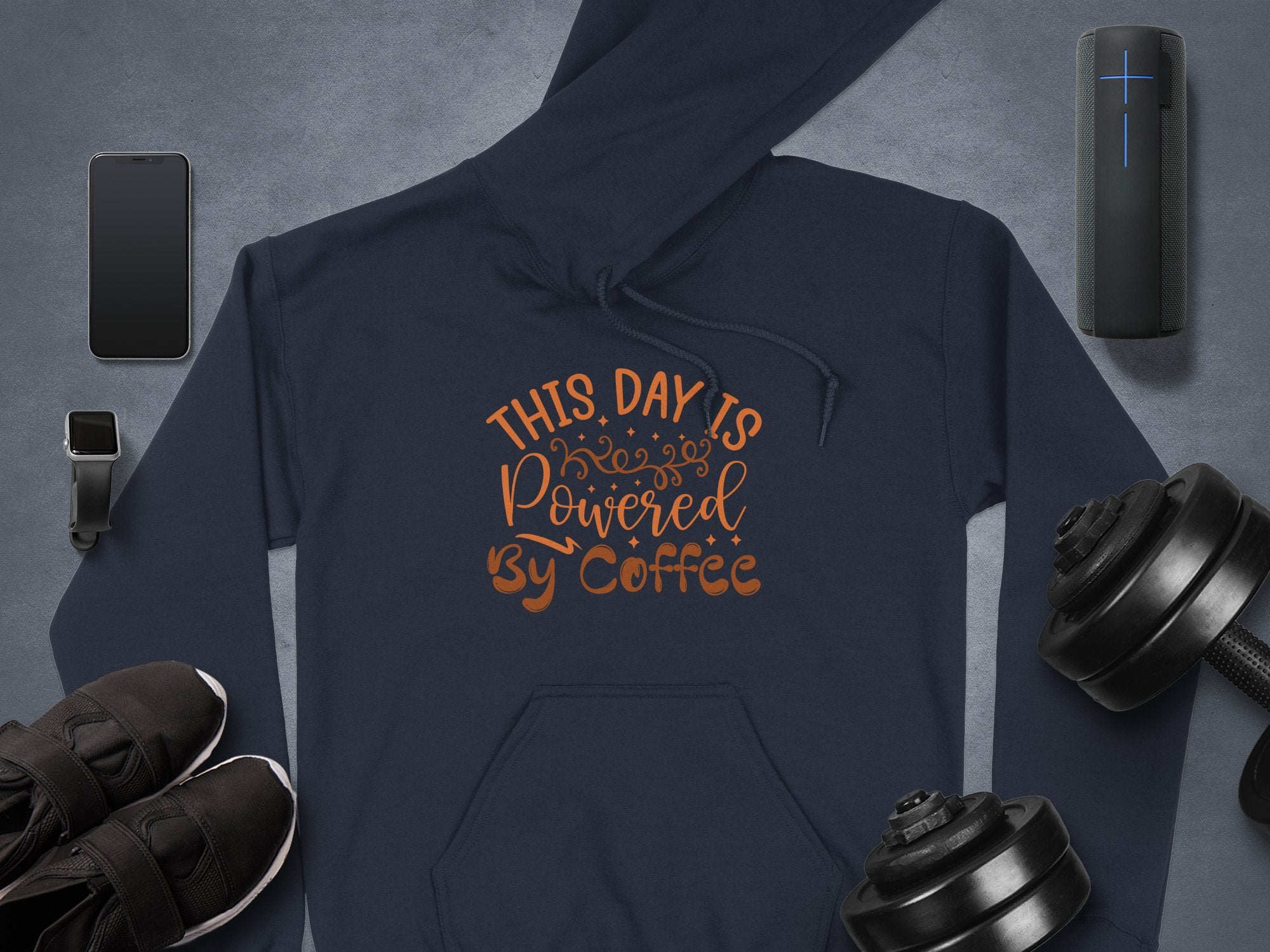 A flat lay features a stylish navy hoodie from Garment Graphics with This Day is Powered by Coffee in brown lettering, ideal for coffee lovers. Its surrounded by dumbbells, a smartwatch, smartphone, black sneakers, and a portable Bluetooth speaker on a gray backdrop.