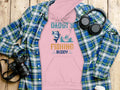 A pink Garment Graphics hoodie featuring Daddys Fishing Buddy and a fishing cartoon is displayed on wood, flanked by a blue plaid shirt, headphones, a camera, and a white coffee cup lid—ideal for outdoor adventures and cozy days.