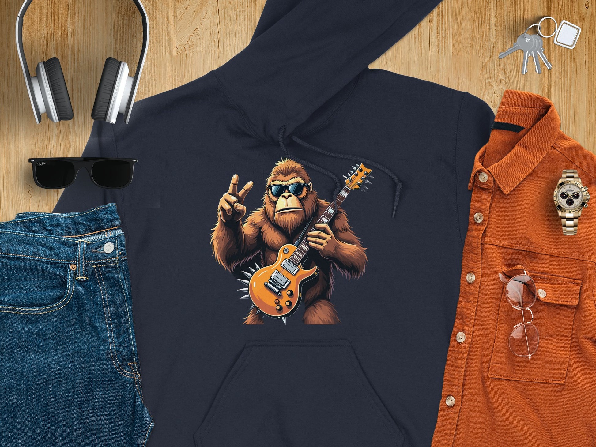 Flat lay of Garment Graphics cozy navy Guitar Playing Gorilla Hoodie, perfect for stylish music and animal lovers. It features a gorilla with a guitar and peace sign, surrounded by jeans, headphones, sunglasses, a watch, keys, and an orange button-up shirt.