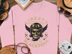 A pink sweatshirt by Garment Graphics, with Liberty Motorcycles Specialist Blackpool 2023 and a skull wearing goggles, showcases a biker vibe. Its surrounded by sandals, a camera, a straw hat, and a notebook on a wooden surface.