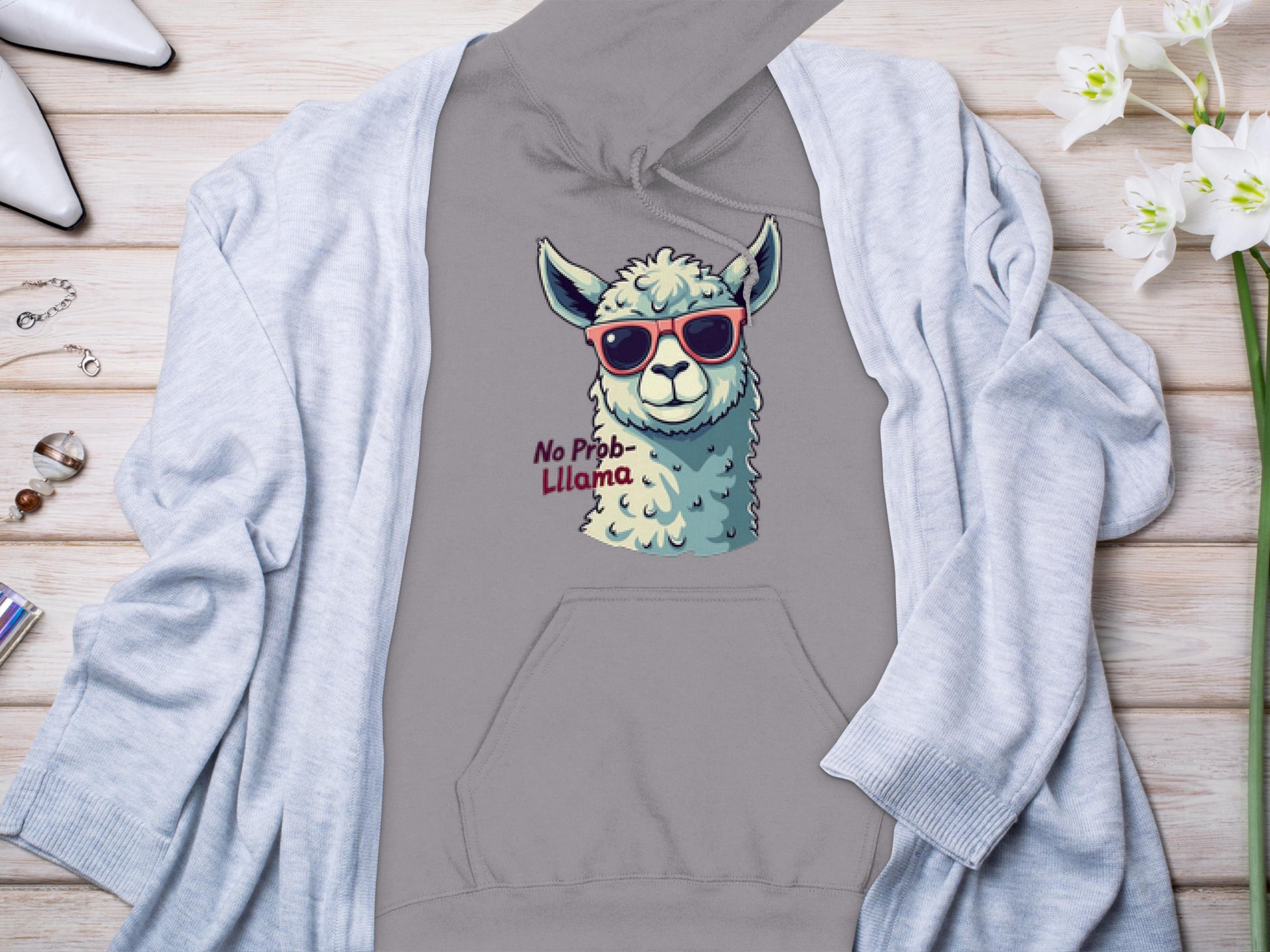 The Garment Graphics unisex gray hoodie, featuring a cartoon llama in sunglasses and No Prob-Llama caption, lays on wood. Its surrounded by a light gray cardigan, white pointy shoes, a watch, rings, and delicate white flowers.