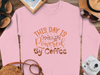 Garment Graphics pink graphic sweatshirt features a playful This Day is Powered by Coffee design in vibrant orange. Its styled on wood alongside a notebook, straw hat, camera, sunglasses, and sandals—ideal for an inspired day out.