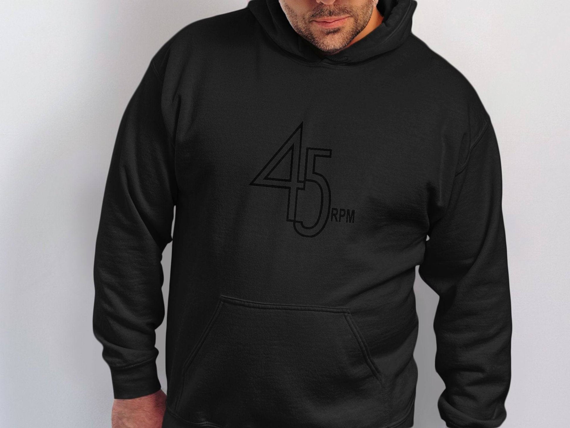 Someone is wearing a black Retro Music Hoodie by Garment Graphics, featuring 45 RPM stylishly printed on it. Set against a plain white background, their face is partially visible as they keep their hands warm in the pocket—a great homage for Vinyl Record Lovers.