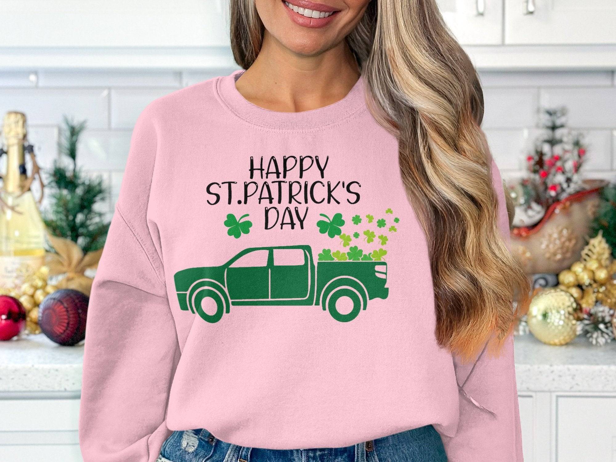 Sporting a pink Garment Graphics festive sweatshirt with a green truck, shamrocks, and Happy St. Patricks Day, one stands by shelves filled with holiday ornaments and a small Christmas tree.