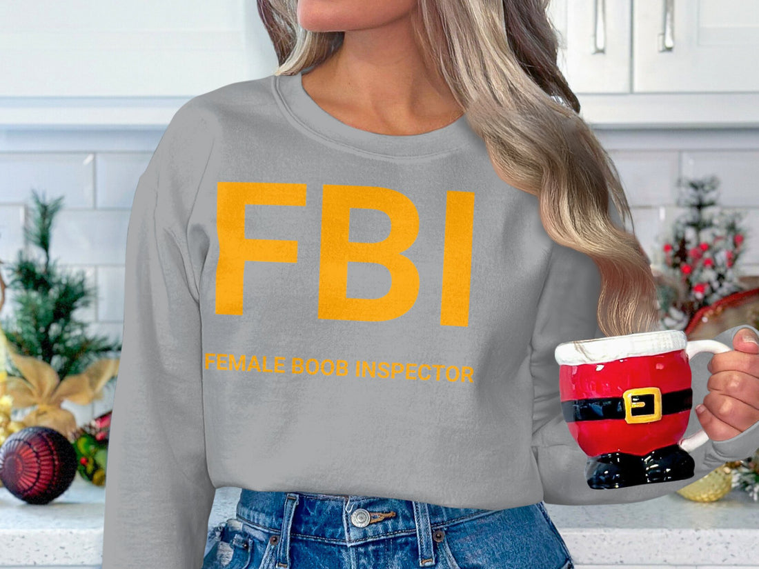 Someone wearing a Garment Graphics white unisex graphic sweatshirt stands outside. The sweatshirt displays large yellow FBI letters with smaller text Female Boob Inspector, against a blurred greenery and track backdrop.