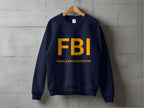 A navy Garment Graphics unisex sweatshirt hangs on a wooden hanger against a gray wall. FBI is printed in large yellow letters across the chest, with Female Boob Inspector humorously below.