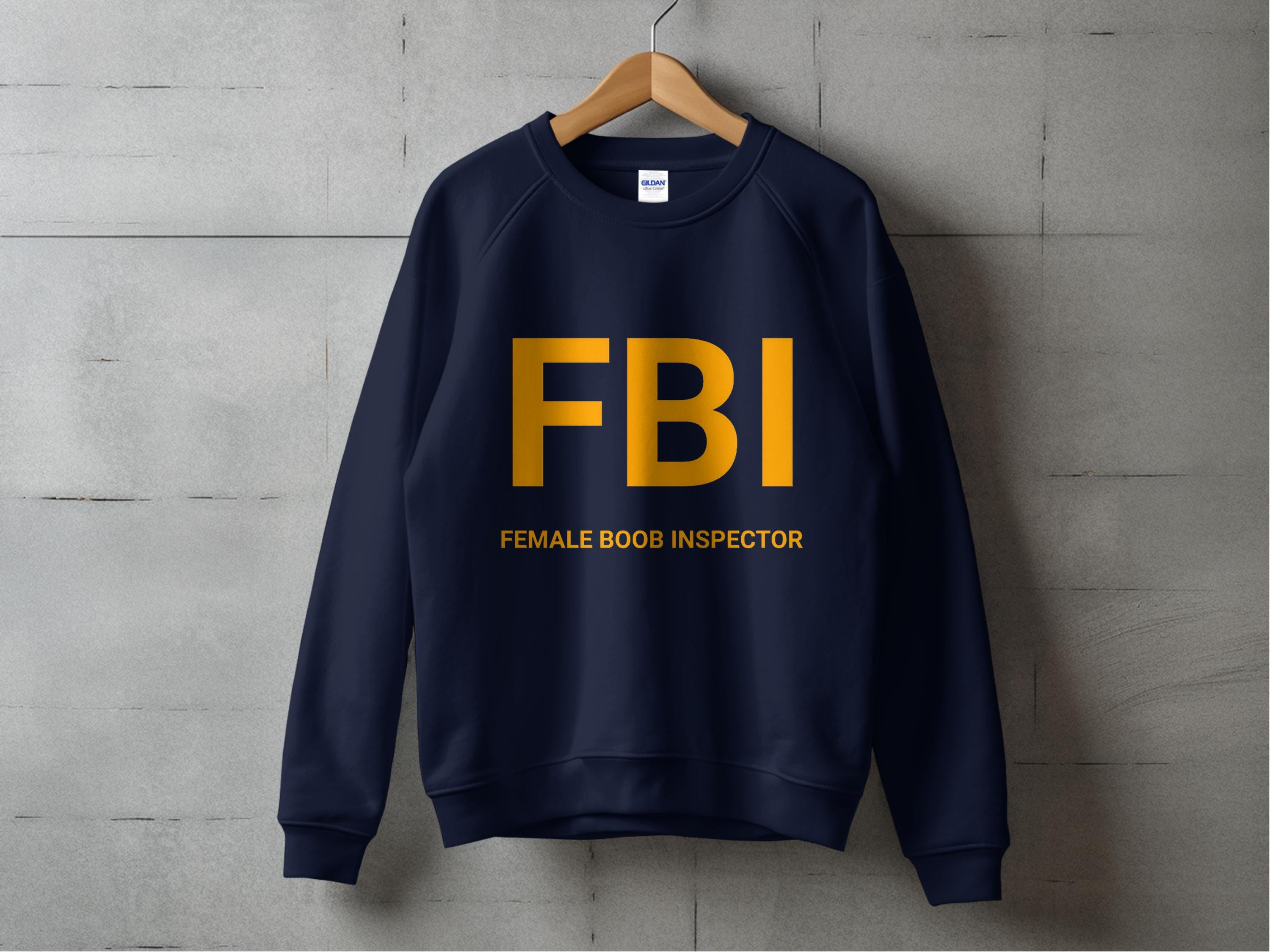 A navy Garment Graphics unisex sweatshirt hangs on a wooden hanger against a gray wall. FBI is printed in large yellow letters across the chest, with Female Boob Inspector humorously below.