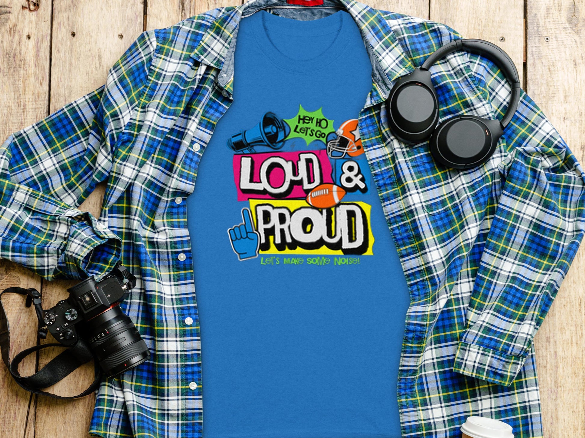 A blue Garment Graphics t-shirt emblazoned with Loud & Proud, Lets Make Some Noise! lies on a wooden surface beside a blue plaid shirt. Ideal for football fans looking to express their passion, it is accompanied by black headphones and a camera.