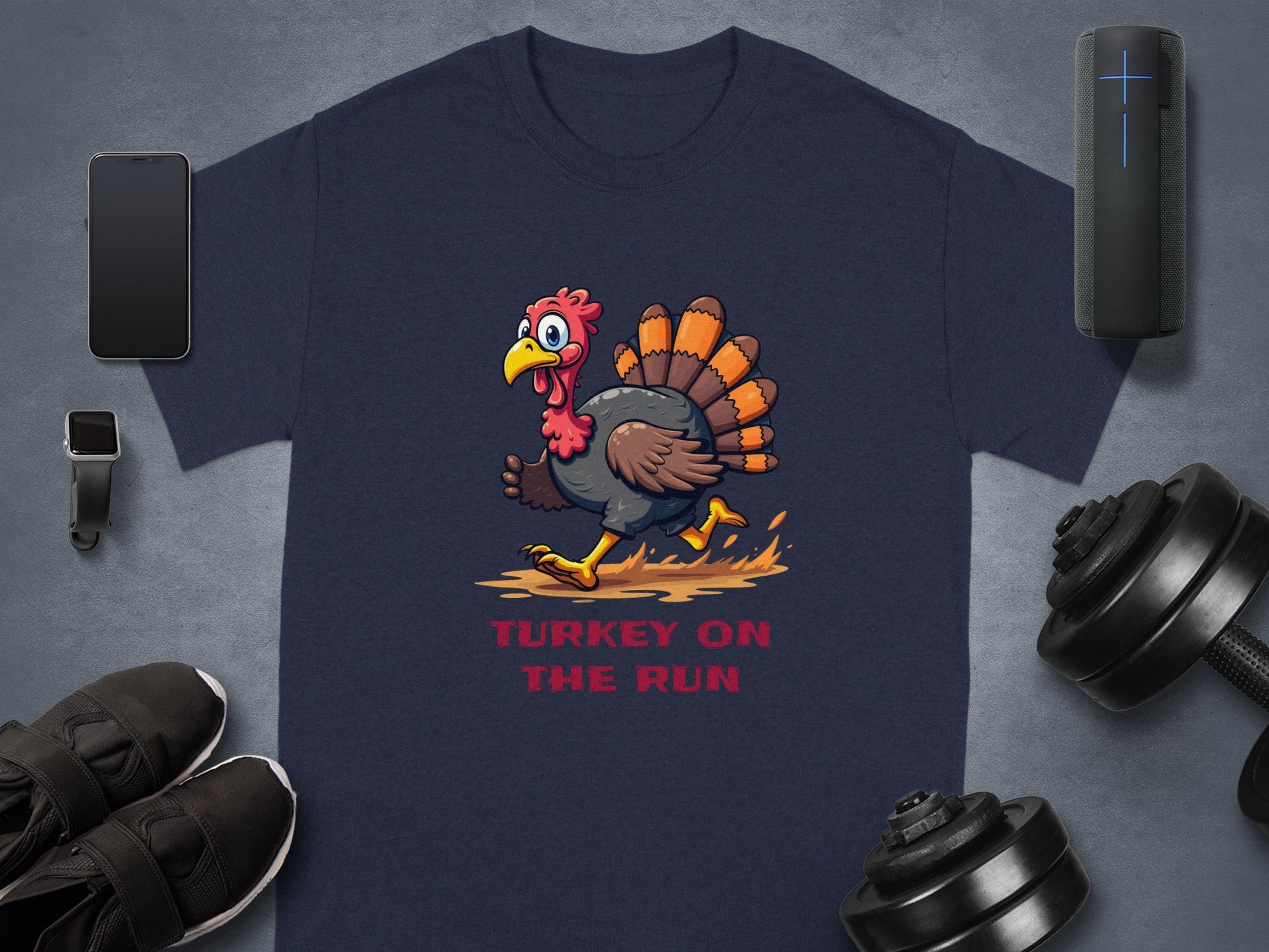 A navy blue Garment Graphics T-shirt with a classic fit features a cartoon turkey running with Turkey on the Run text. It is displayed on a gray surface surrounded by a smartphone, smartwatch, Bluetooth speaker, black shoes, and adjustable dumbbells.